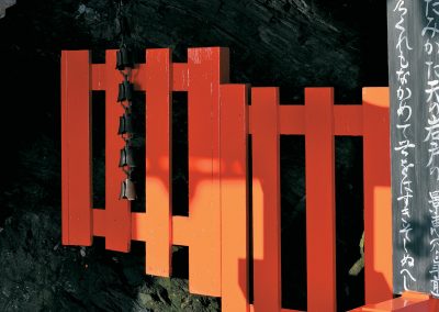 Portail orange sur le fond noir des rochers cachés dans les ombres de l'arrière-plan. Une guirlande de clochettes pend ddevant, des idéogrammes peints verticalement bordent le côté droit de l'image.